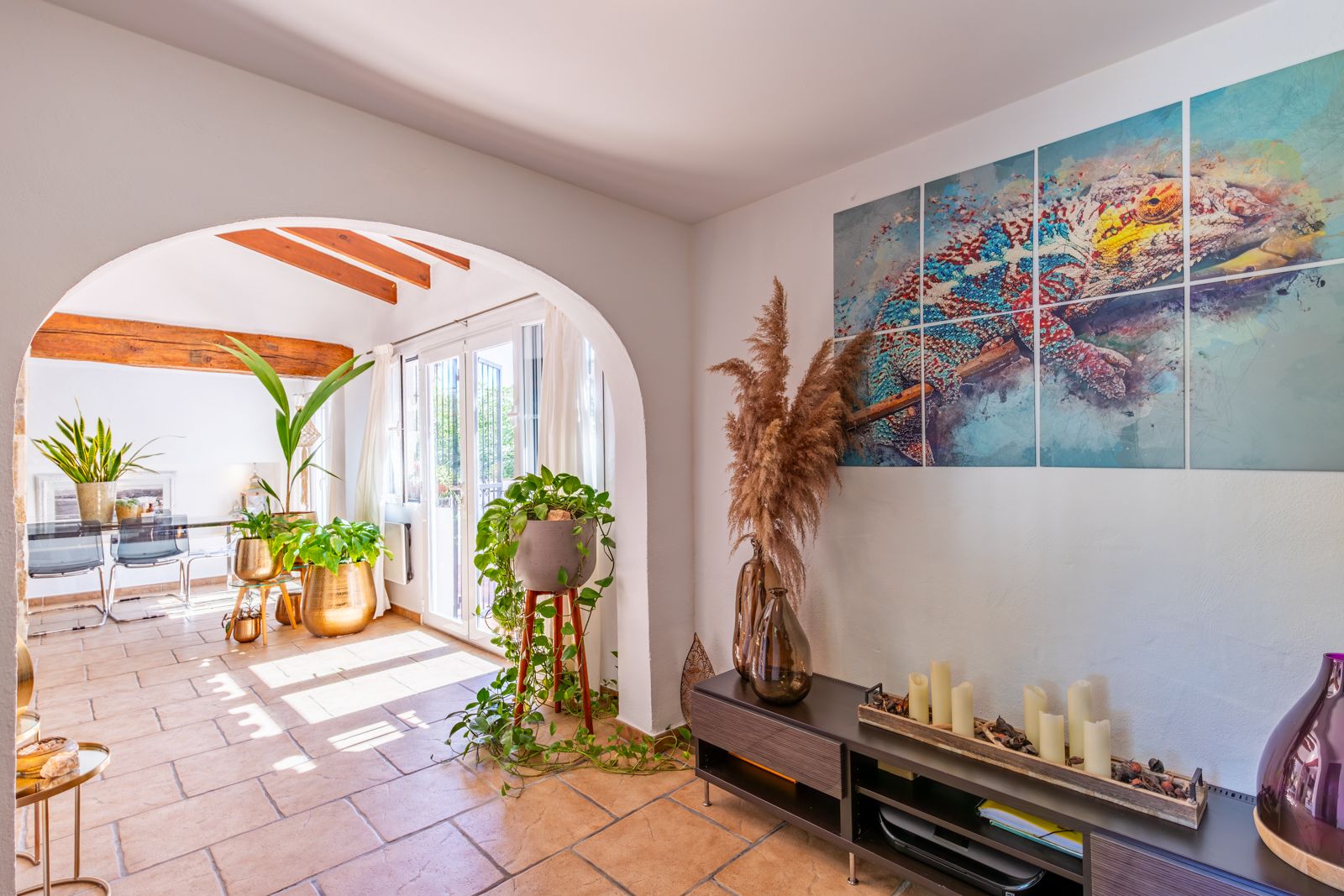 Open-concept living area featuring a Mediterranean arch, terracotta flooring, and exposed wooden beams. Large windows provide ample natural light to the interior.