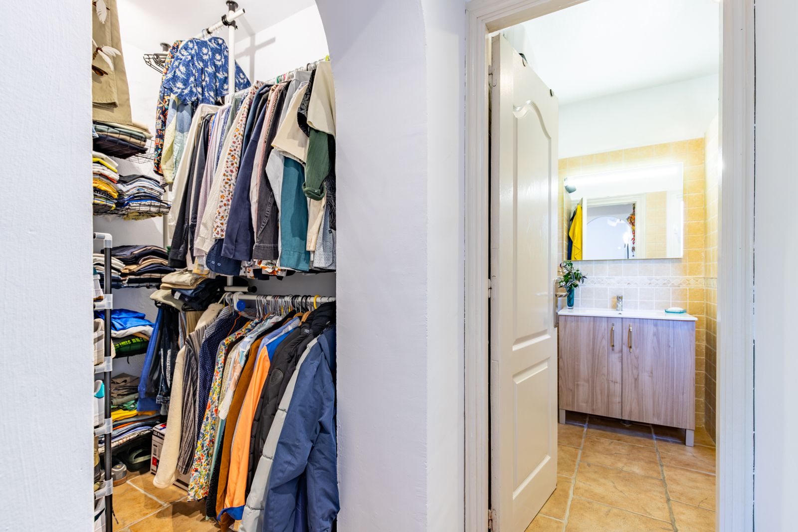 Open walk-in closet with integrated shelving and hanging rails, leading to a bathroom with a wood-finish vanity and backlit mirror.