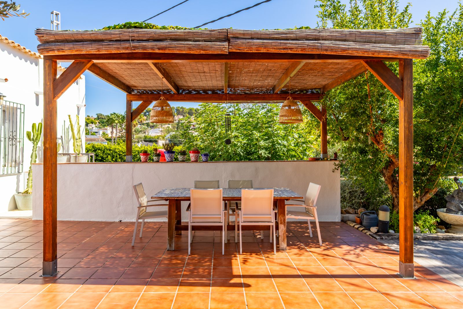 Outdoor dining area featuring a wooden pergola with bamboo roofing, wicker pendant lights, and a tiled table on a terracotta terrace.