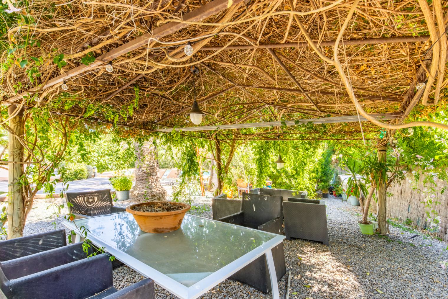 Outdoor dining space shaded by a natural vine-covered pergola, featuring a glass-top table, synthetic rattan seating, and gravel flooring within a lush garden.