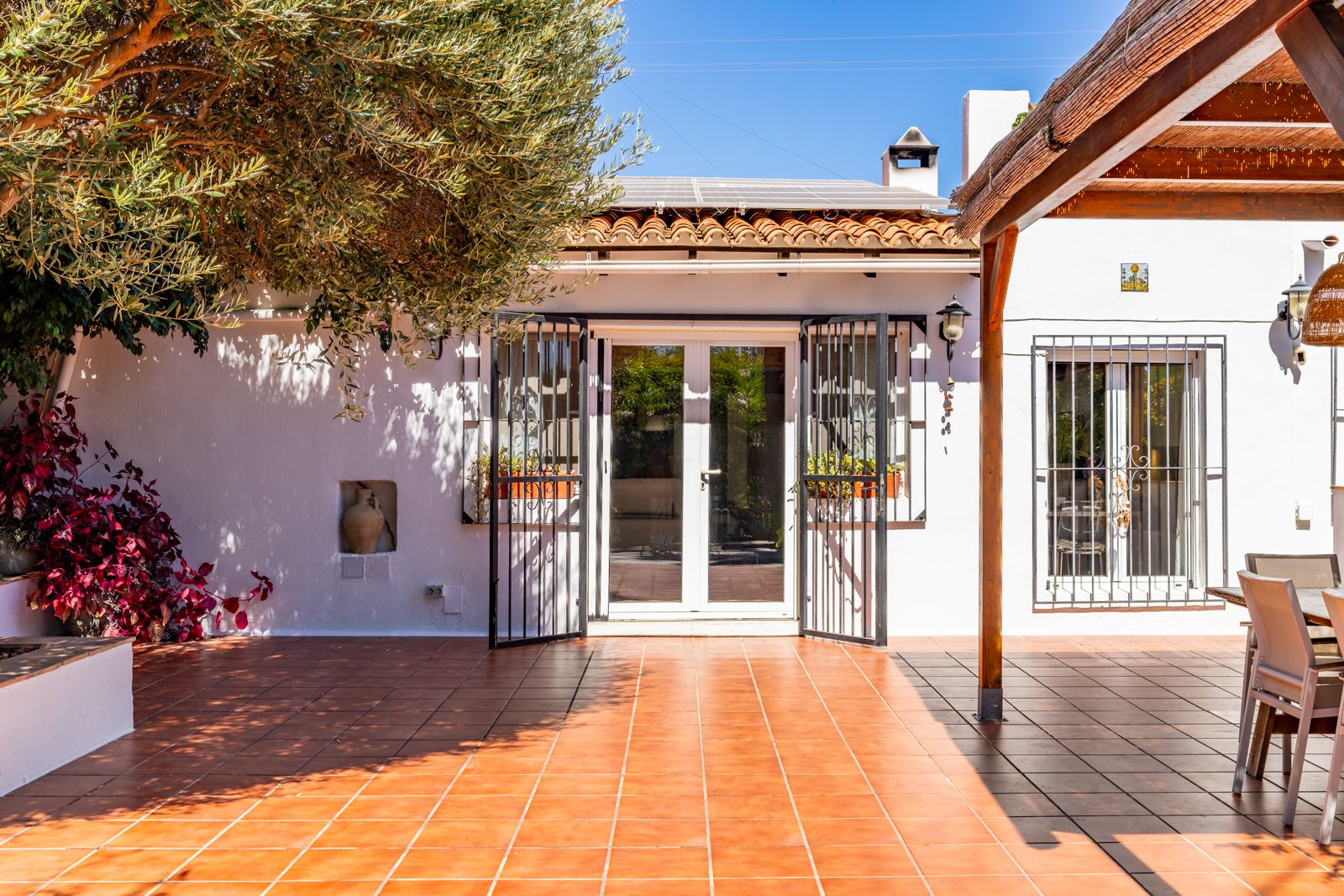 Outdoor terrace with terracotta tiling, a wooden pergola, and rooftop solar panels. The white facade features black ironwork and an olive tree providing natural shade.