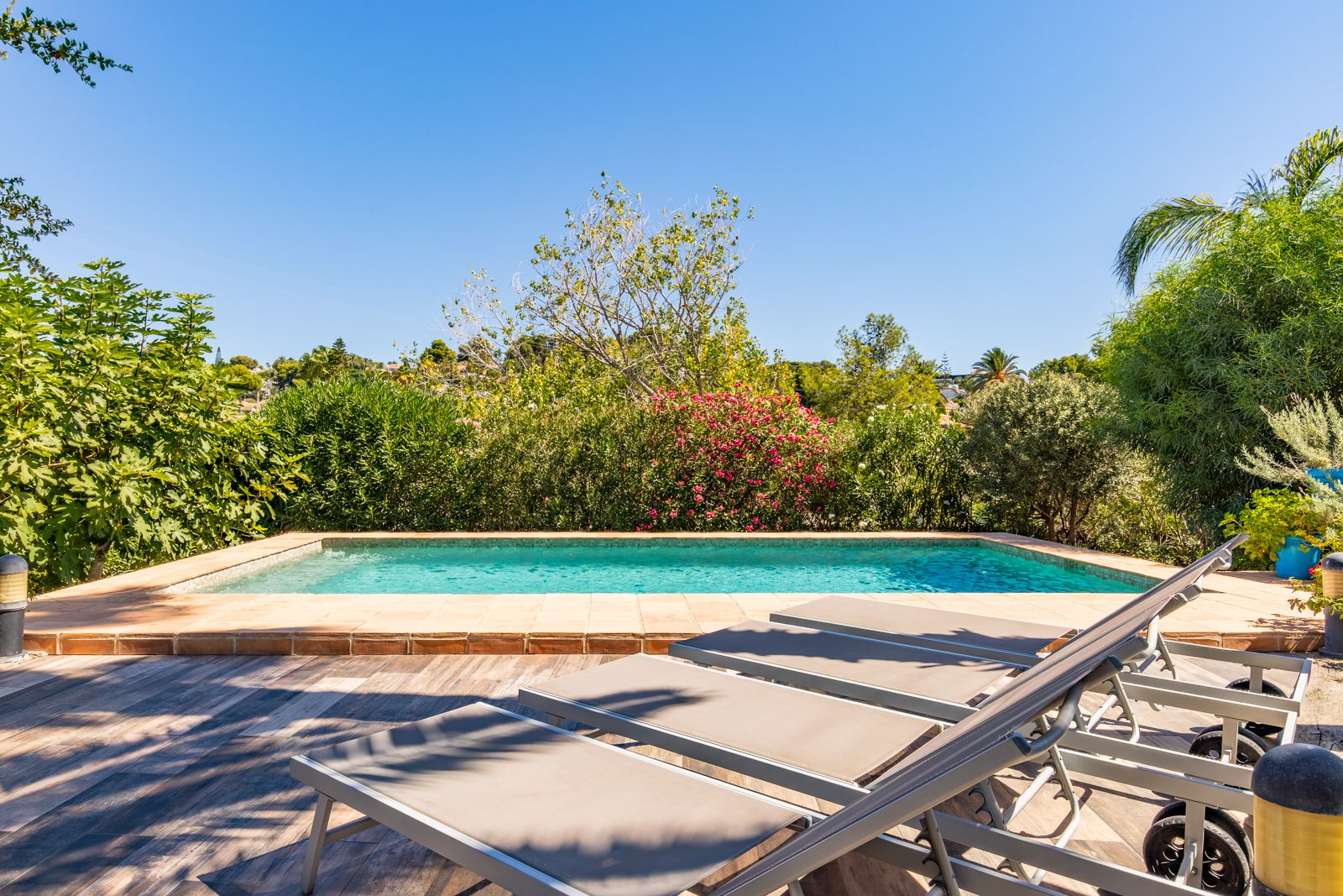 Private swimming pool area featuring a wood-effect tiled terrace, modern sun loungers, and a perimeter of lush Mediterranean greenery.