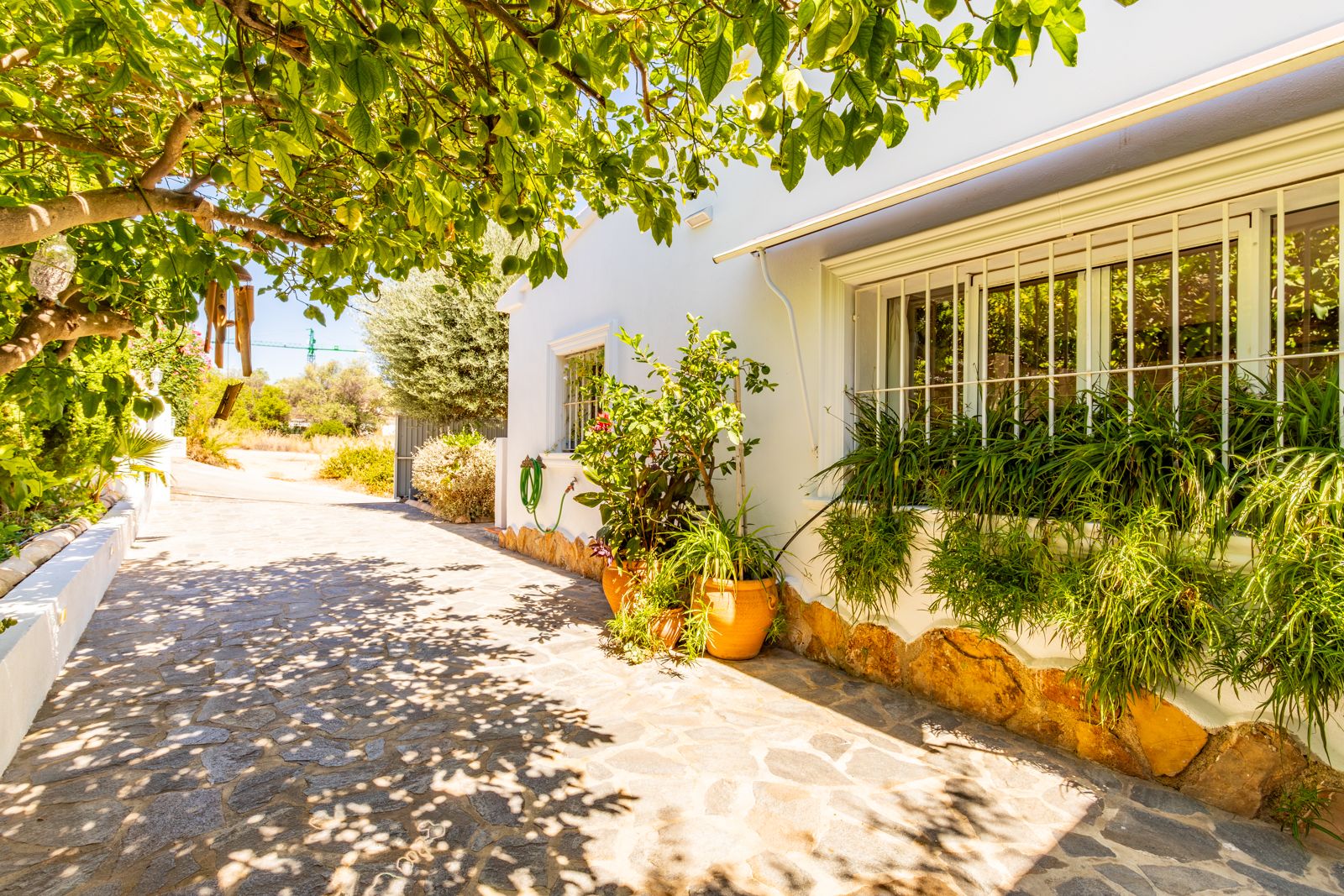 White facade featuring stone accents and barred windows, bordered by lush Mediterranean plants and a natural stone-paved walkway under leafy shade.