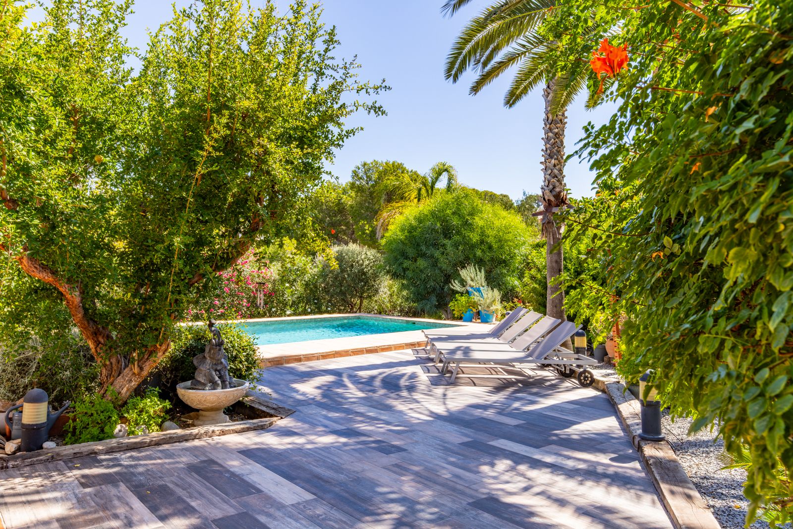 Wood-effect tiled sun terrace featuring grey loungers, a private swimming pool, and lush Mediterranean landscaping with mature trees and palms.