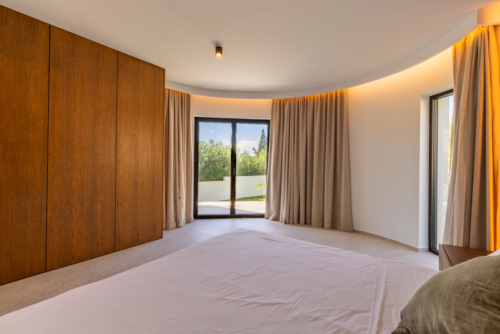 Bedroom featuring a curved wall, custom oak wardrobes, and indirect cove lighting. Large sliding glass doors provide seamless access to the terrace.
