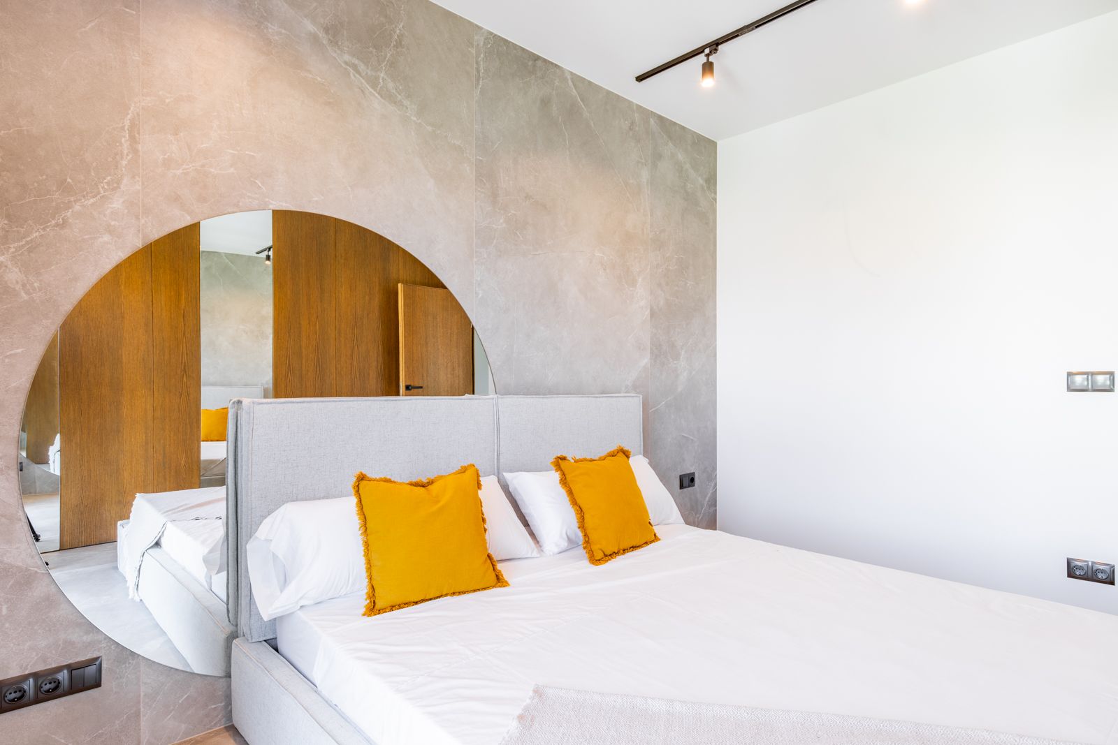 Bedroom featuring a large-format porcelain wall with an integrated circular mirror. Includes a fabric headboard, natural oak paneling, and minimalist track lighting.