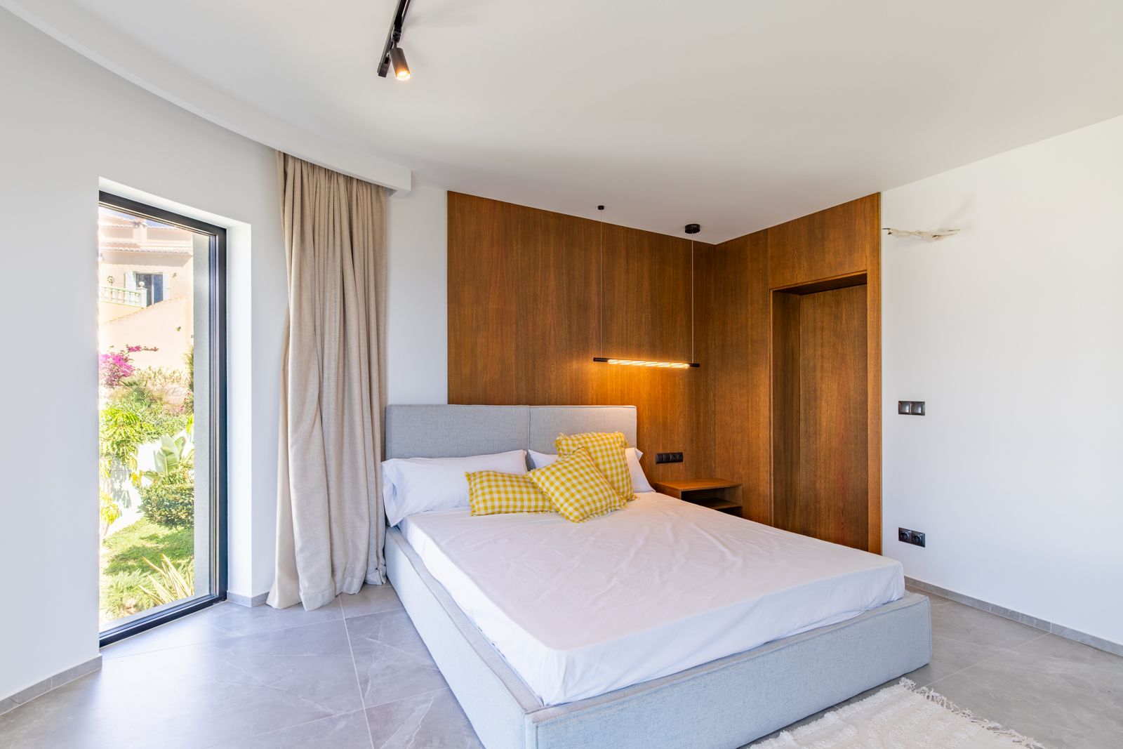 Bedroom featuring natural oak wood paneling, designer pendant lighting, and large-format porcelain floors. A floor-to-ceiling window offers direct views of the garden.