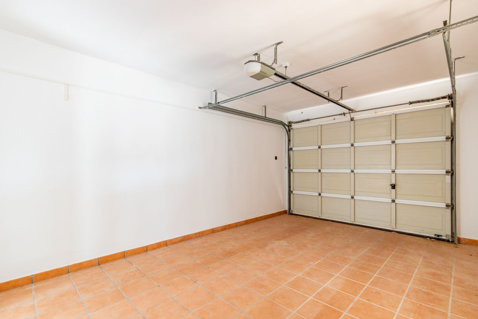 Closed garage featuring terracotta-style ceramic floor tiles, smooth white walls, and a motorized sectional door with a ceiling-mounted automatic opening system.