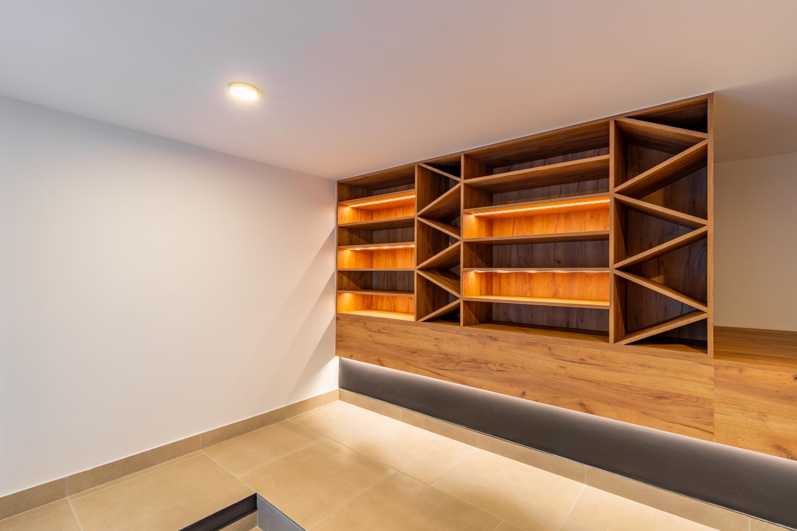 Custom oak wine cellar featuring integrated LED lighting, geometric shelving units, and large-format porcelain flooring in a minimalist setting.