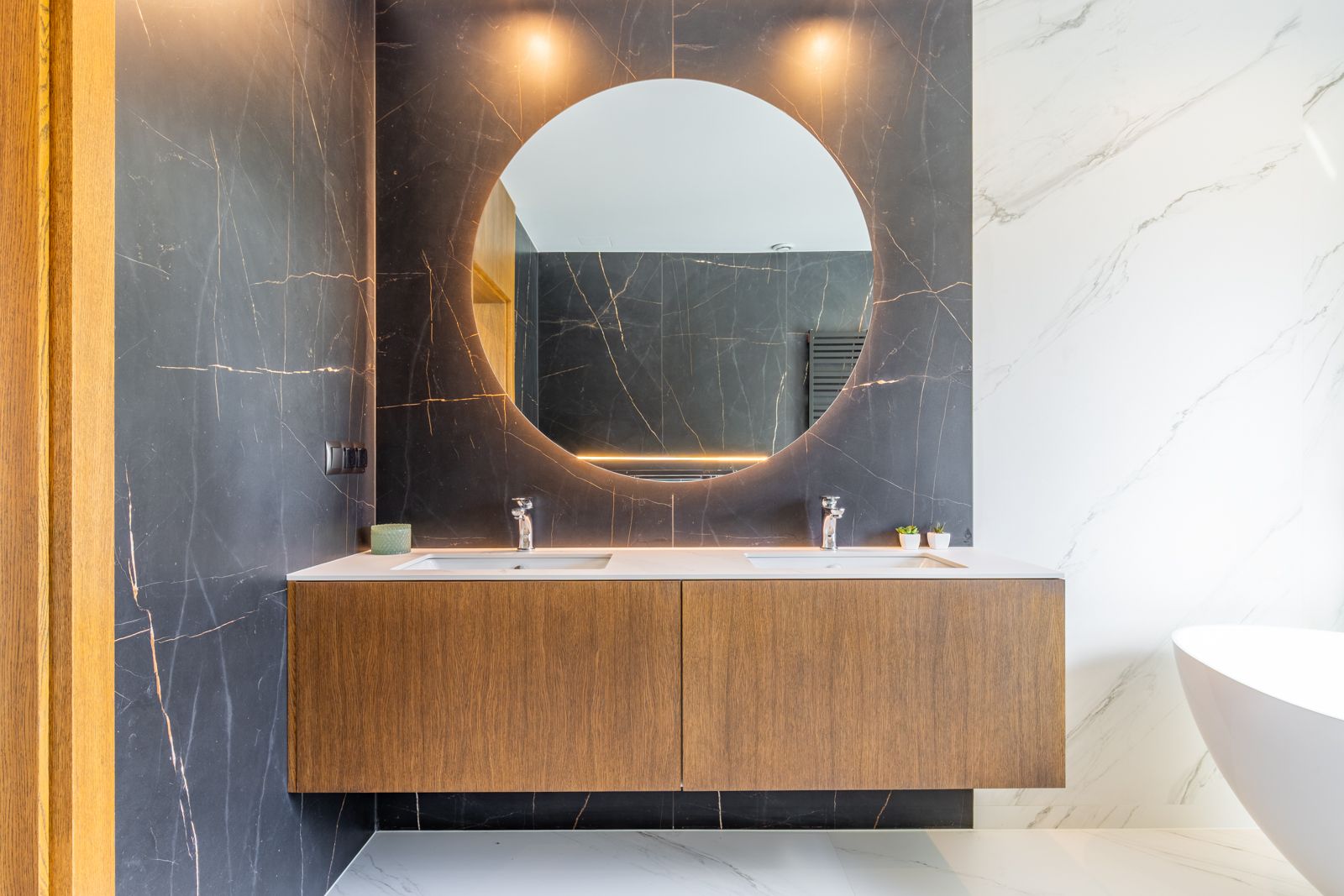 Designer bathroom featuring a floating oak vanity with double sinks, a large backlit circular mirror, and dark porcelain wall tiles with elegant gold veining.