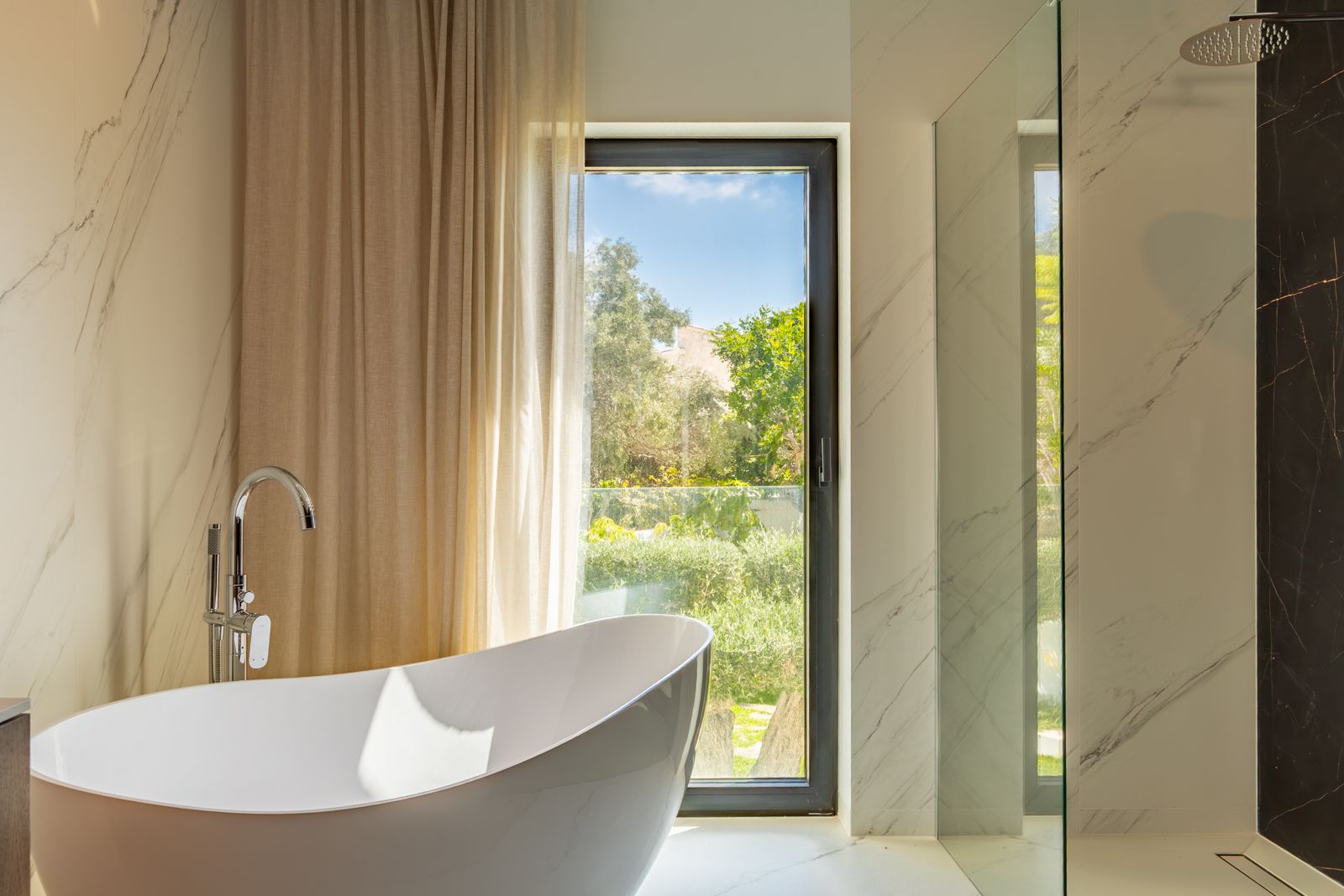Designer bathroom featuring a freestanding bathtub, floor-mounted faucet, and walk-in shower. Marble-effect porcelain walls and a large window offer views of the garden.