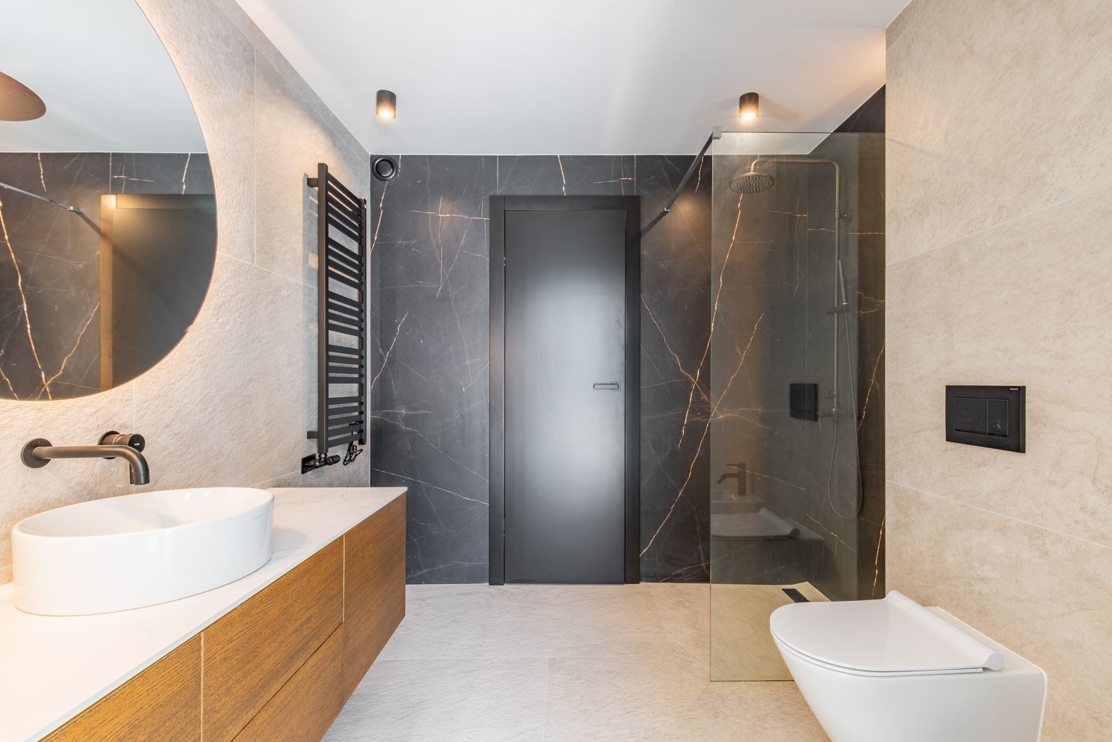 Designer bathroom featuring large-format porcelain tiles, a backlit circular mirror, wooden vanity with vessel sink, and a walk-in shower with matte black fixtures.