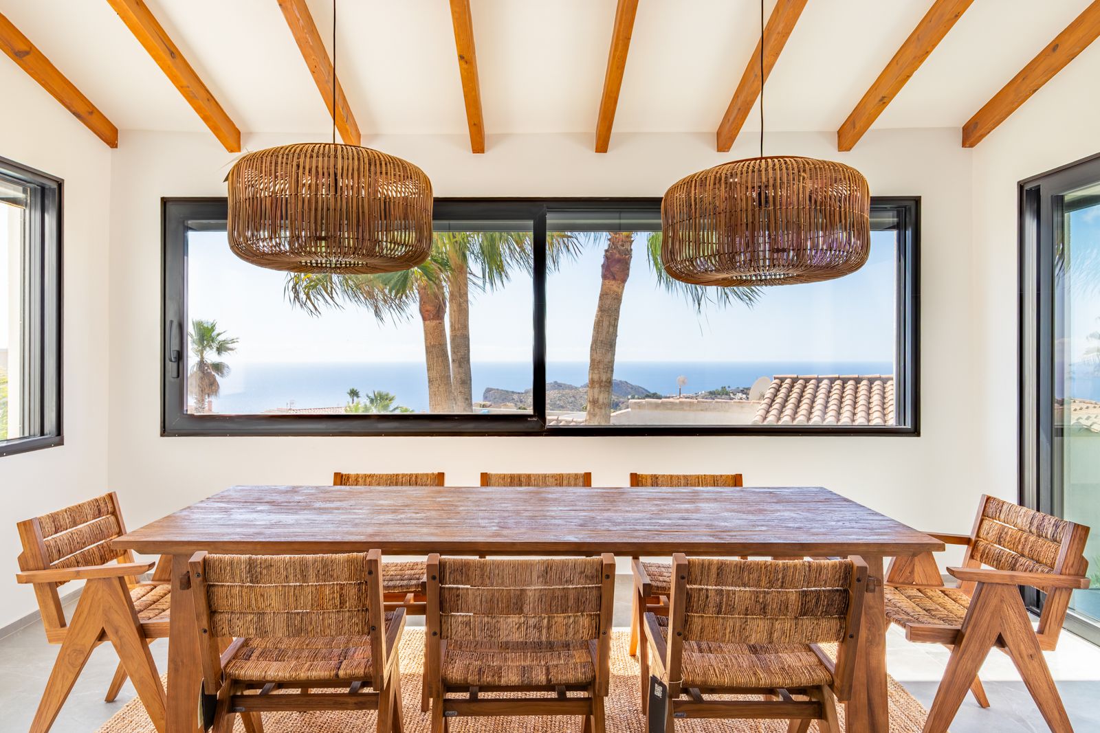 Dining space featuring exposed ceiling beams, a solid wood table, and rattan pendants. A large window frames views of the Mediterranean Sea and palm trees.