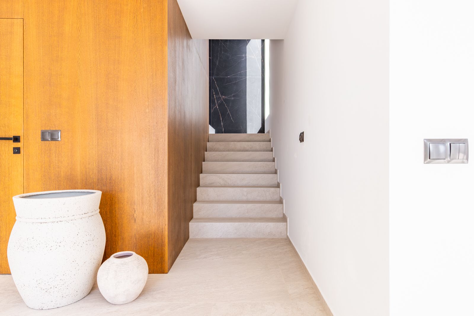 Hallway featuring a porcelain tile staircase, natural oak wood paneling with integrated door, and a black marble-effect feature wall.