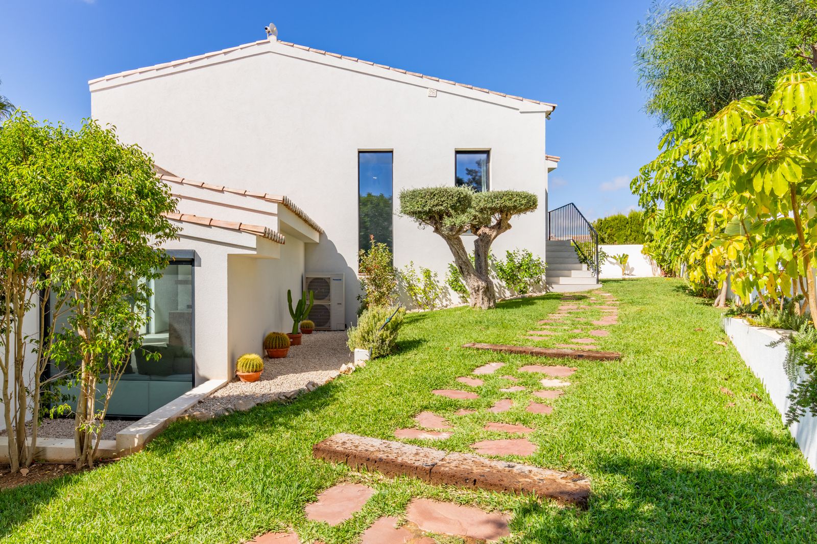 Landscaped Mediterranean garden featuring a manicured lawn, sculptural olive tree, and natural stone path leading to the villa's modern white facade.
