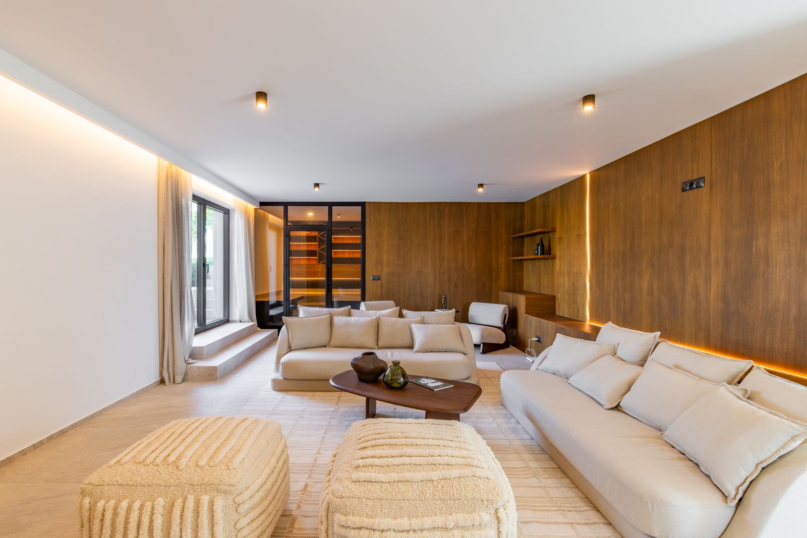 Living area featuring natural oak wall paneling with integrated LED lighting, neutral-toned sofas, and a view towards a glass-enclosed wine cellar.