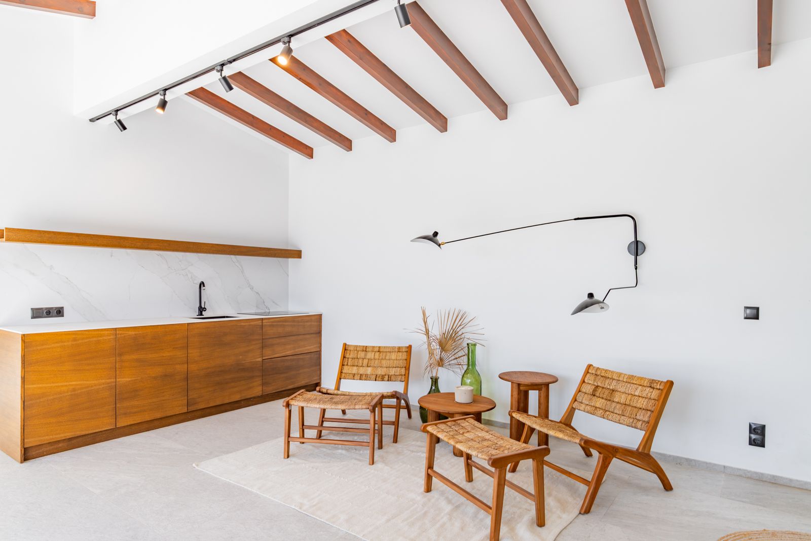 Living space featuring an oak kitchenette with sintered stone surfaces, exposed wooden beams, and designer lounge chairs on a large-format porcelain floor.
