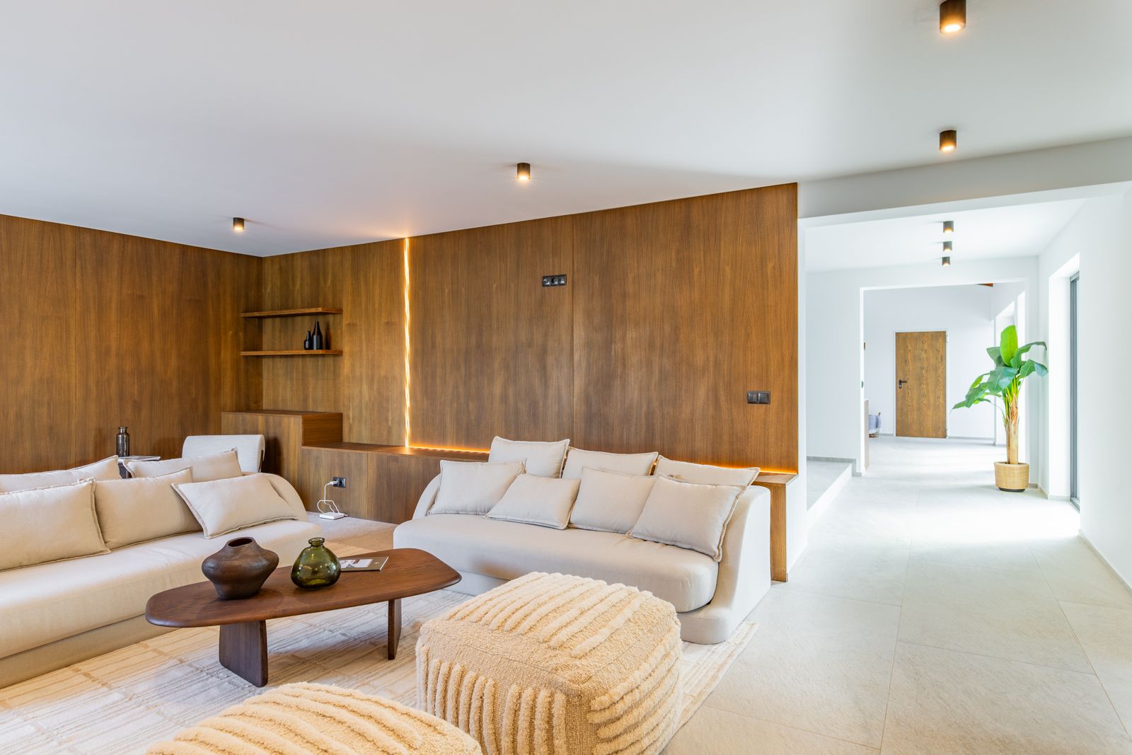 Living space featuring natural oak wall panels with integrated LED lighting, neutral sofas, and large-format porcelain flooring under recessed technical spotlights.