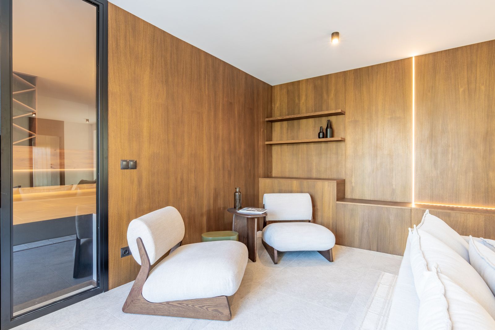 Lounge area featuring natural oak wood paneling, integrated vertical LED lighting, and large-format porcelain flooring. Includes designer upholstered armchairs.