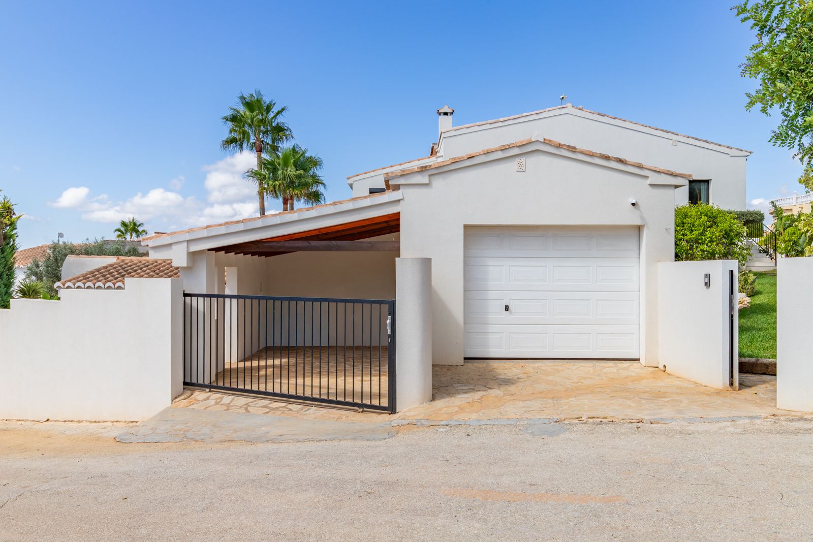 Main entrance featuring a closed garage and a covered carport with wooden beams. Includes natural stone paving, a black metal gate, and clean white Mediterranean architecture.