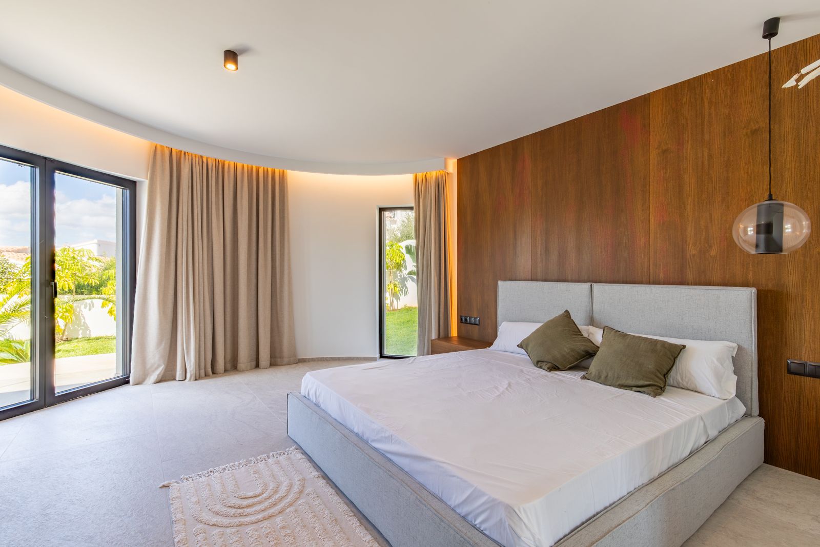 Master bedroom featuring natural oak wall paneling, curved architectural ceiling with indirect lighting, and direct garden access through large glass doors.