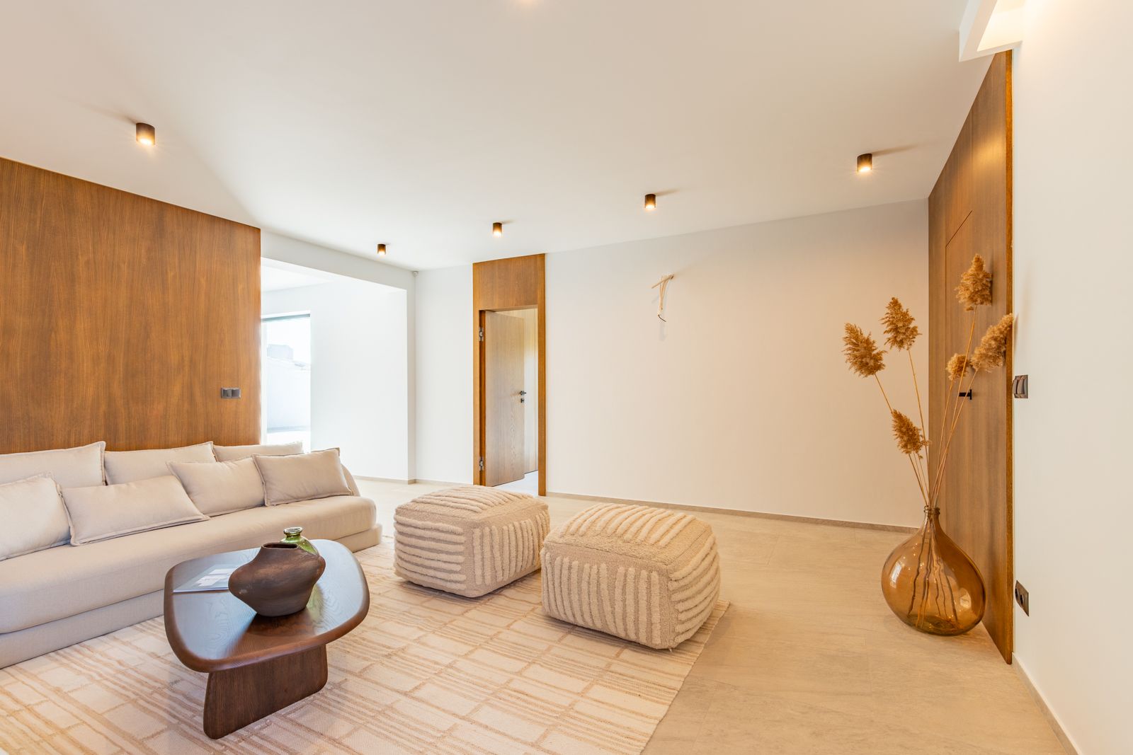 Minimalist living area featuring natural oak wall panels, large-format porcelain flooring, and designer furniture illuminated by recessed ceiling spotlights.