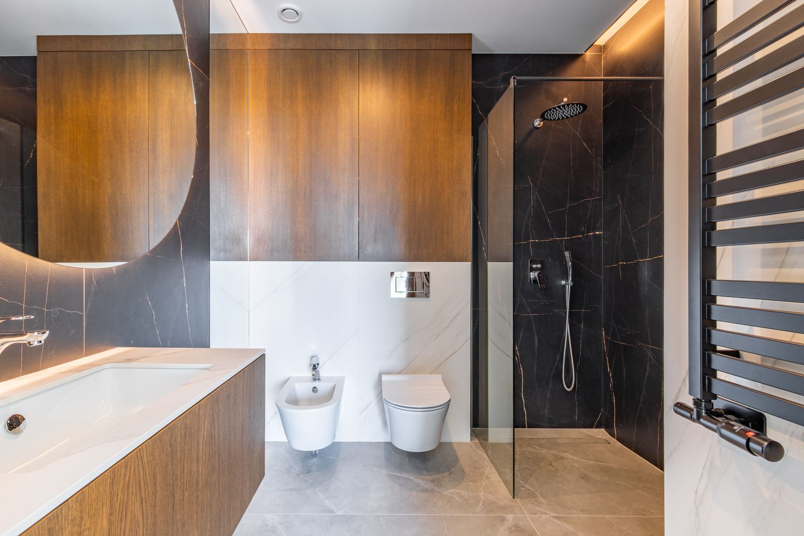 Modern bathroom featuring dark porcelain tiling, a walk-in shower, wall-hung fixtures, and natural oak cabinetry beneath a large backlit circular mirror.