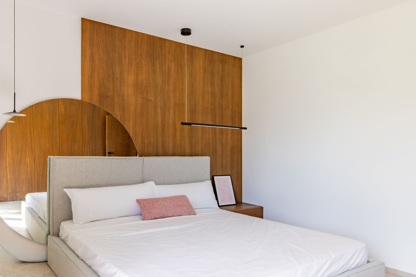 Modern bedroom featuring natural oak wall paneling, a grey fabric headboard, and a large circular mirror. Minimalist black pendant lighting and an integrated wooden nightstand.