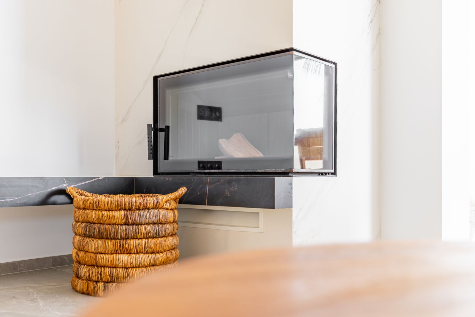 Modern corner fireplace with glass enclosure, integrated into white sintered stone cladding with a black marble ledge and a natural fiber log basket.