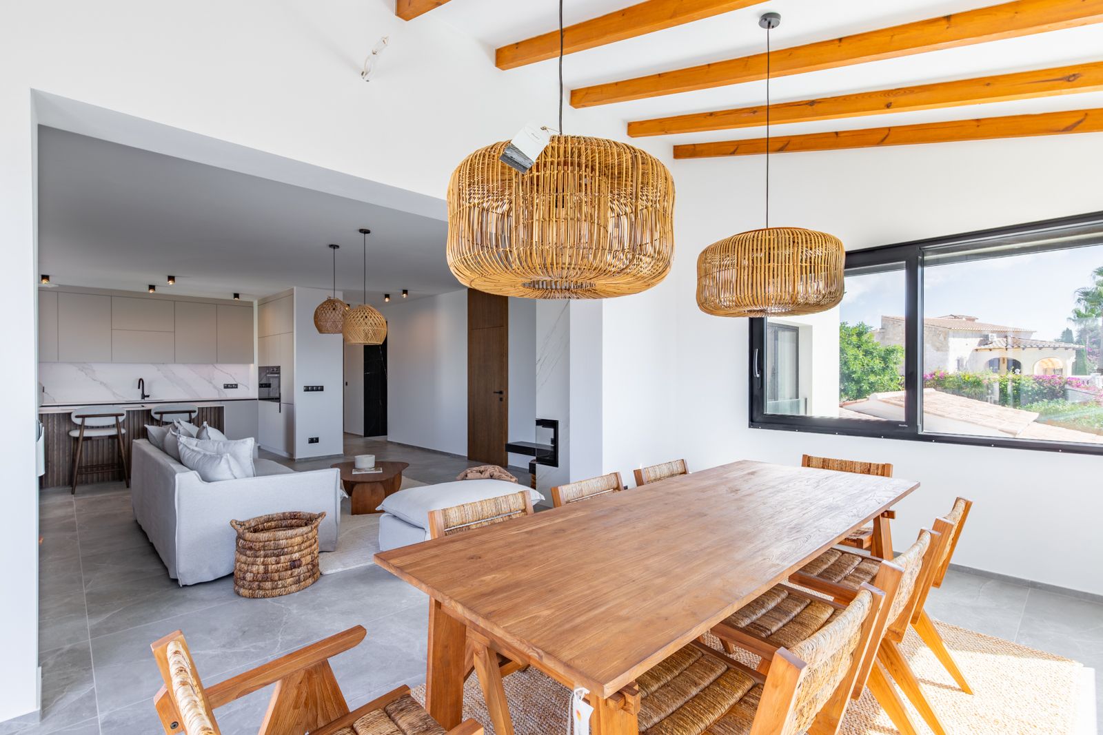 Open-concept dining area featuring a solid wood table, exposed ceiling beams, and rattan pendants, seamlessly connected to the living room and kitchen.