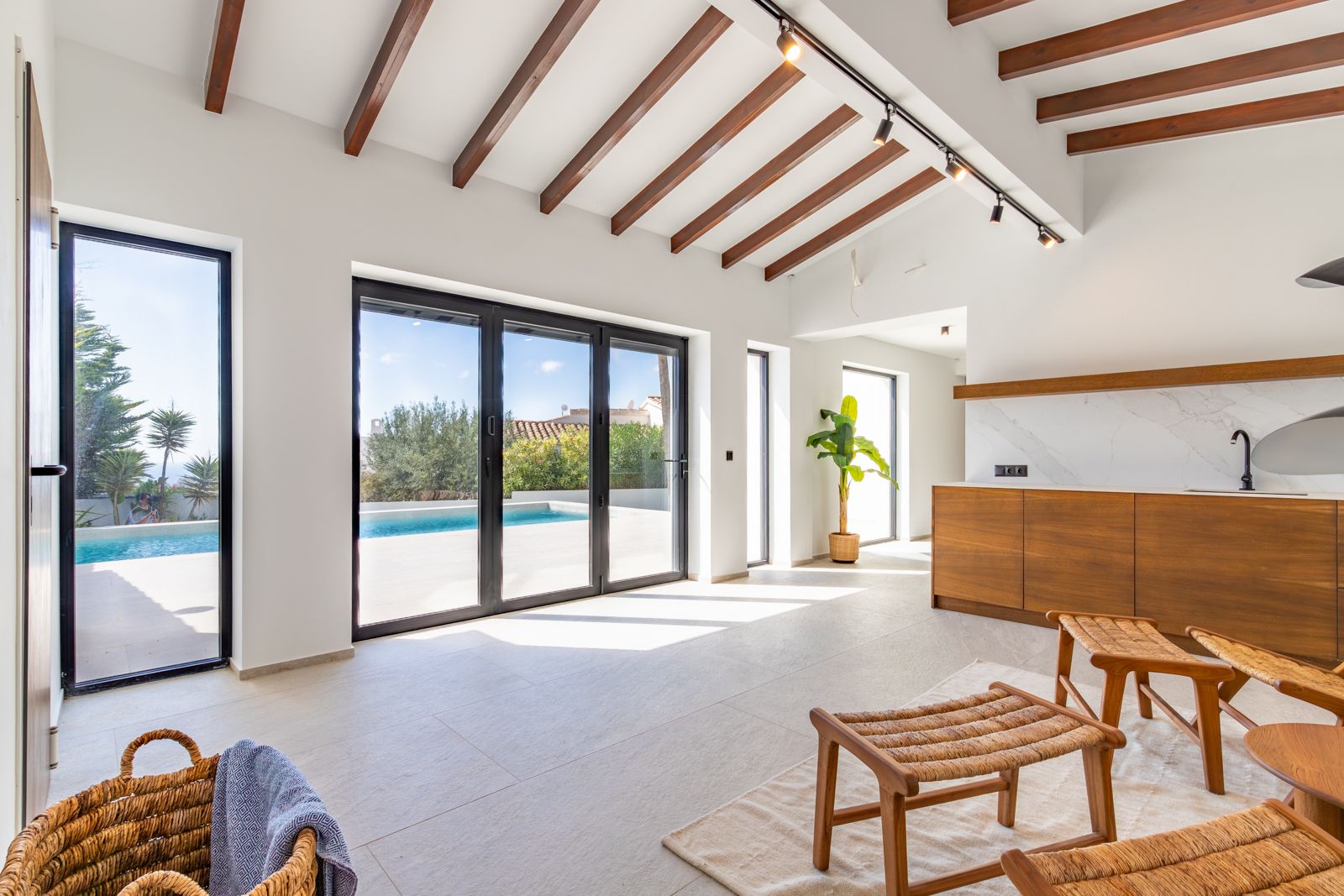 Open-plan living space featuring exposed wooden beams, large-format porcelain flooring, and floor-to-ceiling windows offering direct access to the pool area.