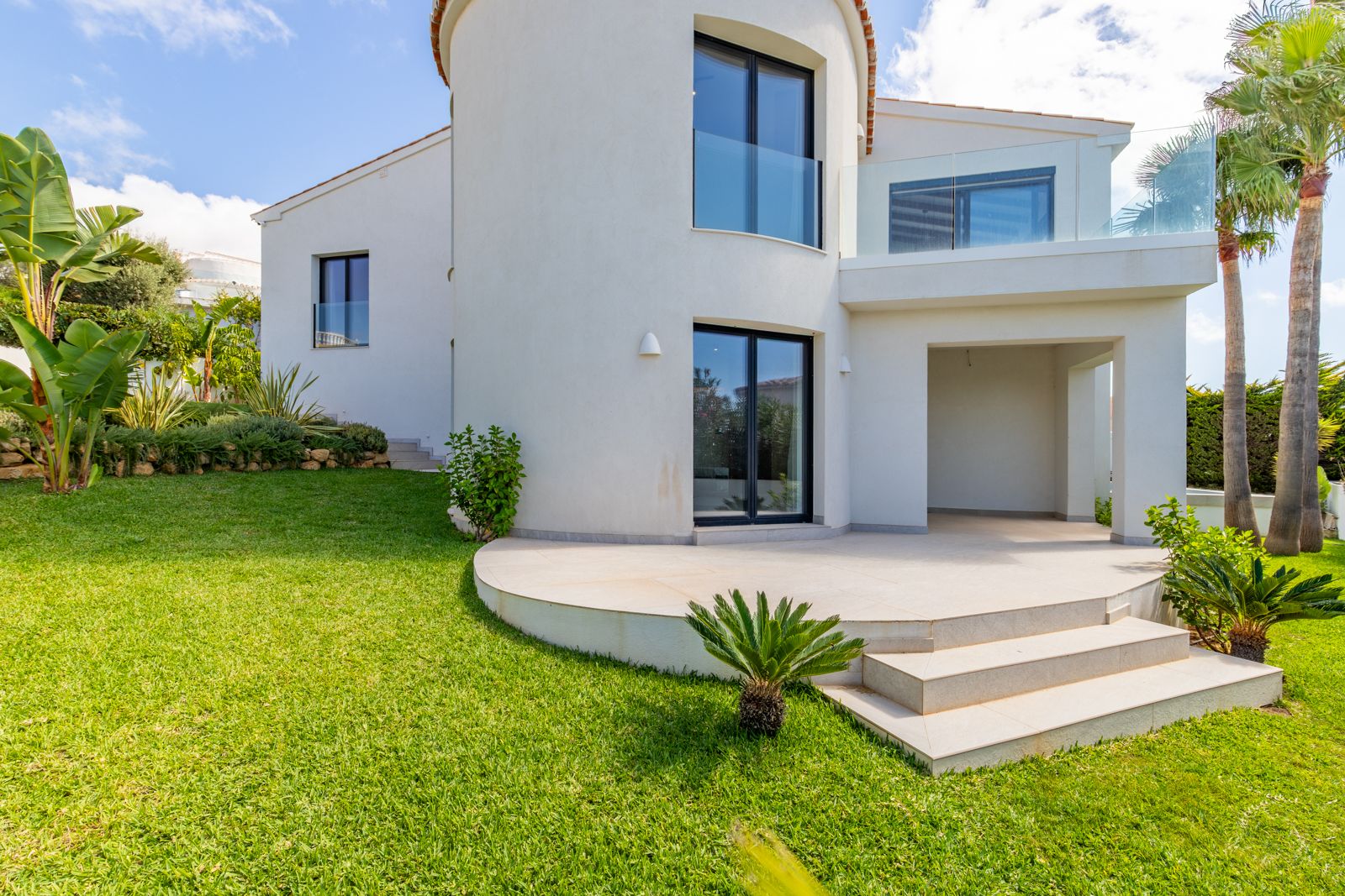 Rear facade featuring a curved architectural tower, dark aluminum window frames, and glass-railed balconies. The manicured lawn is bordered by palm trees and tropical plants.