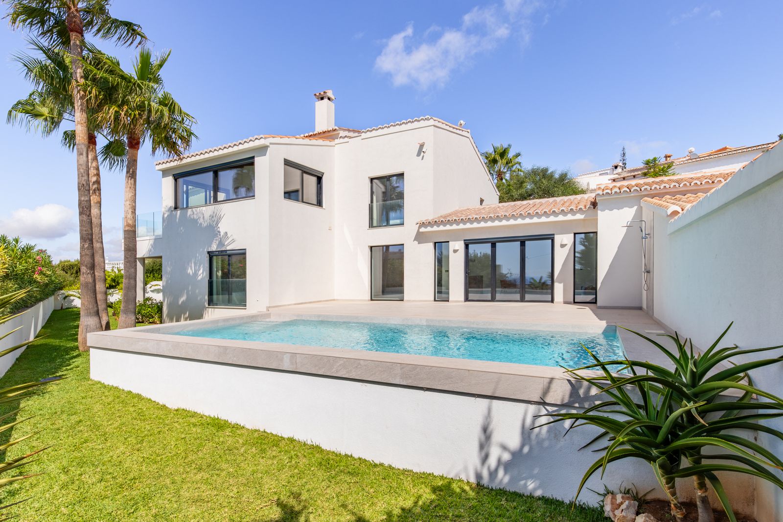 Rear facade of the villa featuring a saltwater pool, porcelain tile terrace, and triple-glazed windows. Landscaped garden with lawn and mature palm trees.