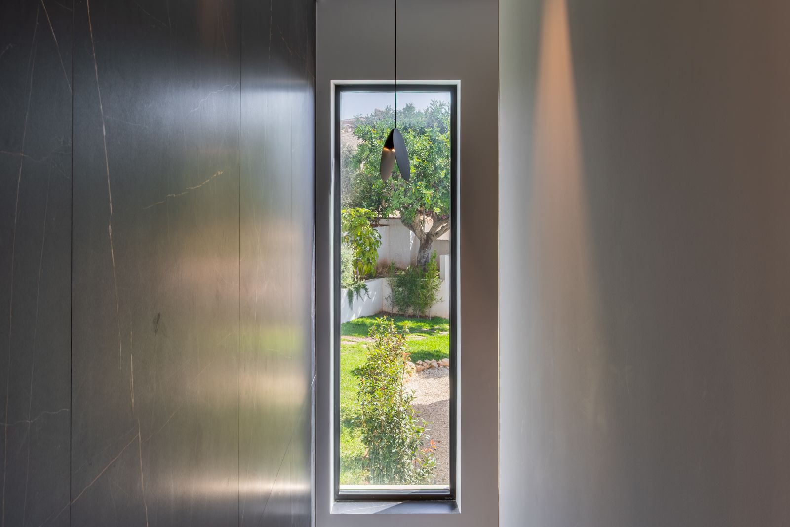 Vertical triple-glazed window framing the garden view, flanked by dark large-format porcelain wall panels and a minimalist designer pendant light.