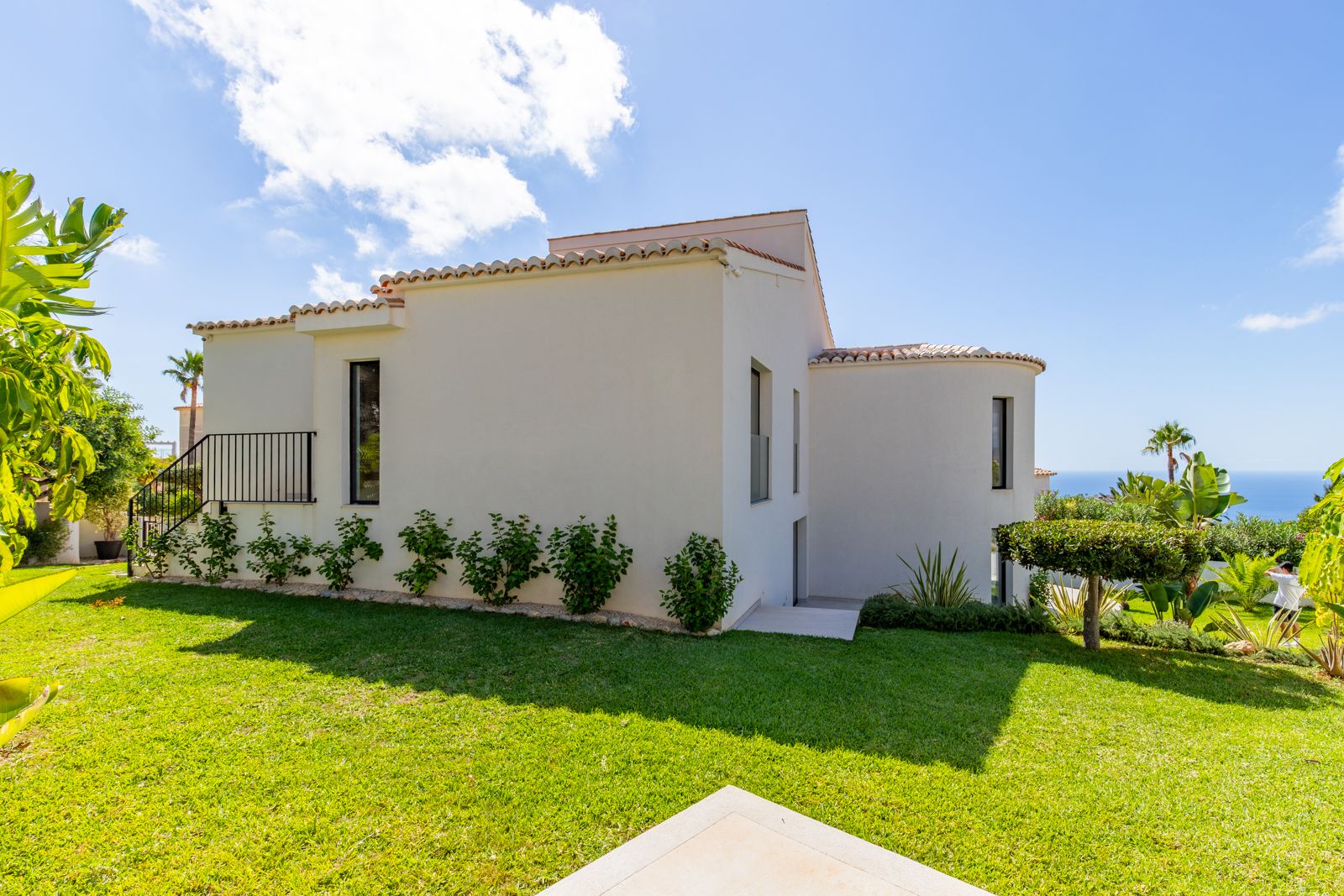 White Mediterranean facade with traditional roof tiles, a manicured lawn, and sea views framed by tropical plants and clear blue skies.