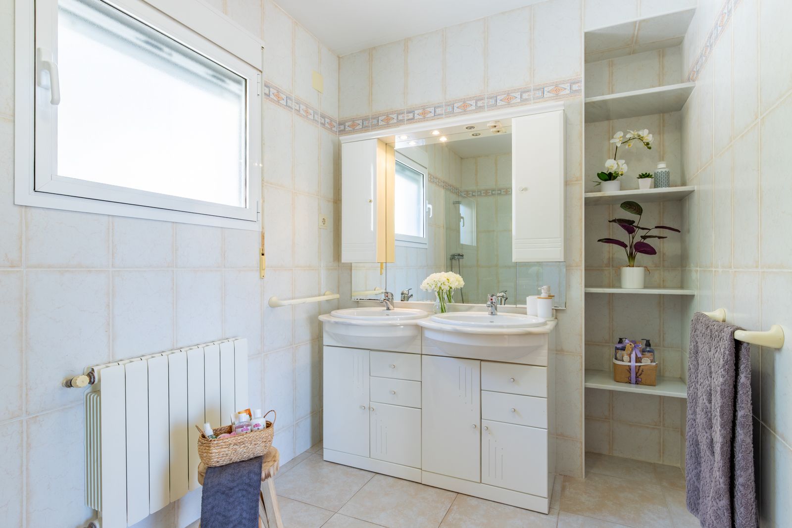 Bathroom featuring a double vanity with white cabinetry, integrated mirror lighting, and built-in shelving. Tiled walls with a decorative border and a wall-mounted radiator.