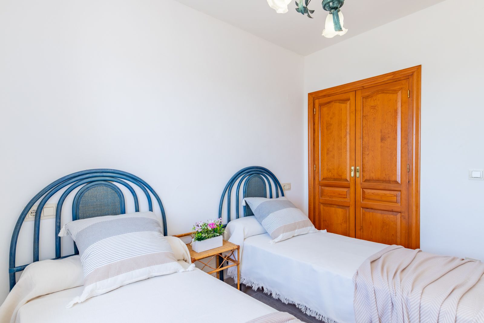 Bedroom featuring twin beds with blue wicker headboards and a built-in natural wood wardrobe. The space offers bright white walls and a functional, clean layout.