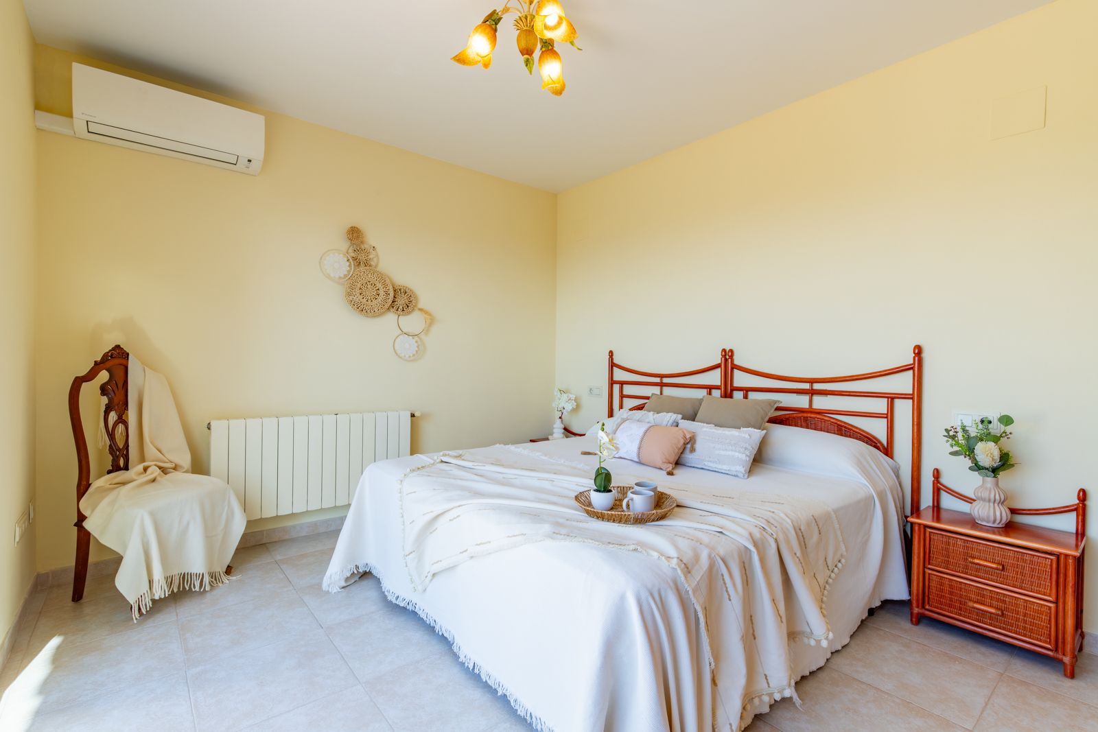 Bright bedroom featuring a double bed with a carved wooden headboard, matching nightstand, split air conditioning, radiator, and neutral tiled flooring.