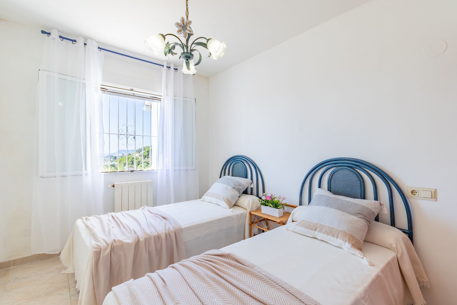 Bright bedroom featuring twin beds with blue arched headboards, central heating, and a window offering natural light and views of the surrounding hills.