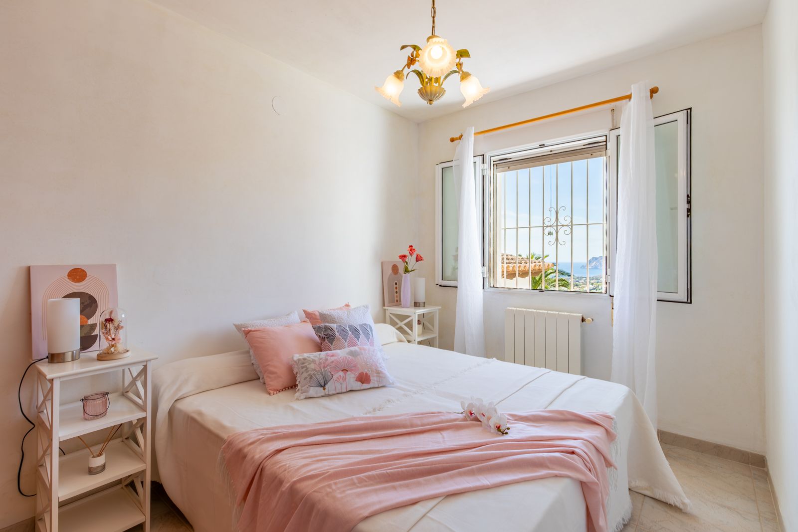 Bright bedroom featuring white walls, tiled flooring, and a double bed. The window provides natural light and views of the Mediterranean Sea and the Peñón de Ifach.