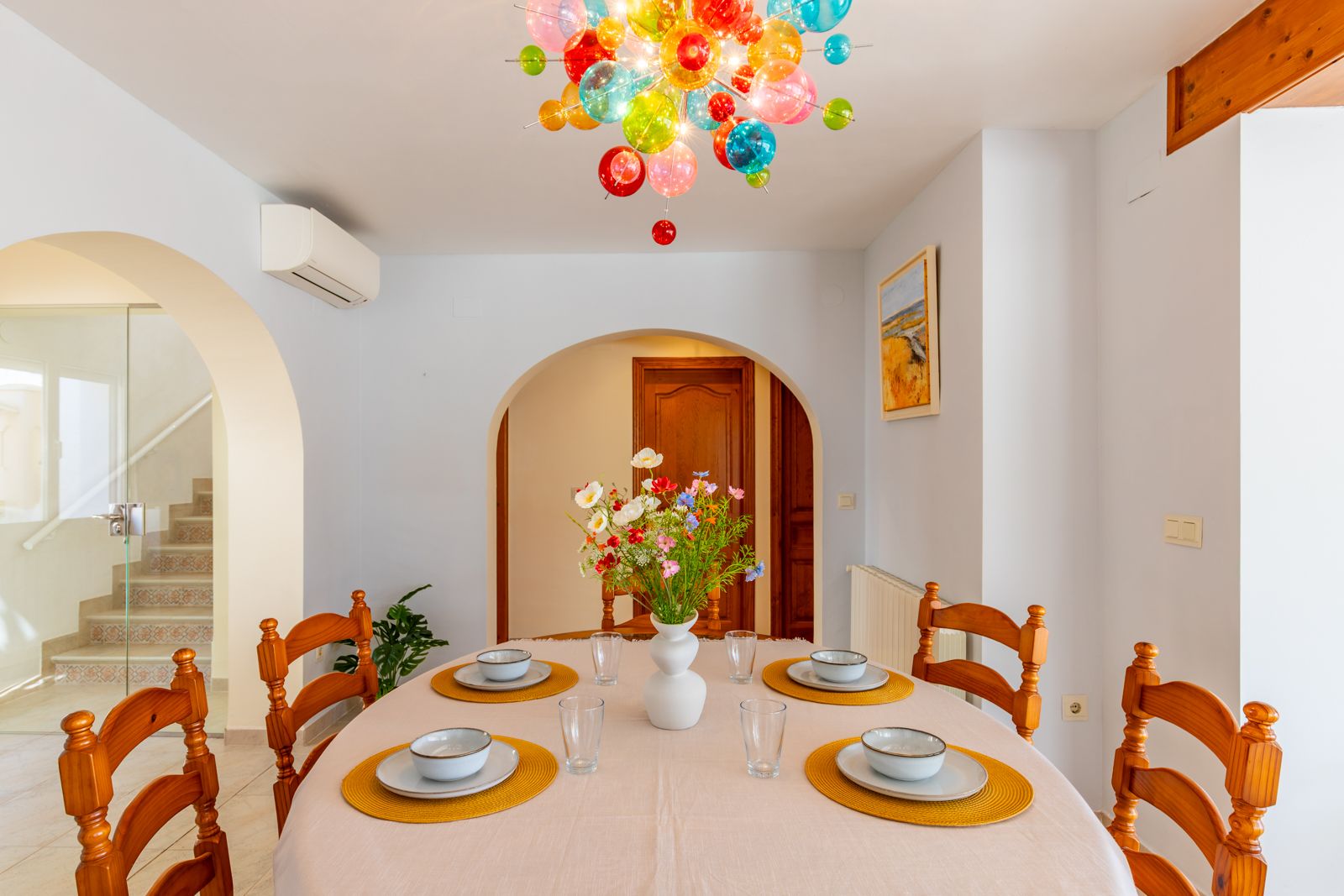 Bright dining area featuring an oval table, wooden chairs, and a vibrant glass bubble chandelier. The space includes traditional arched doorways and a view of the tiled staircase.