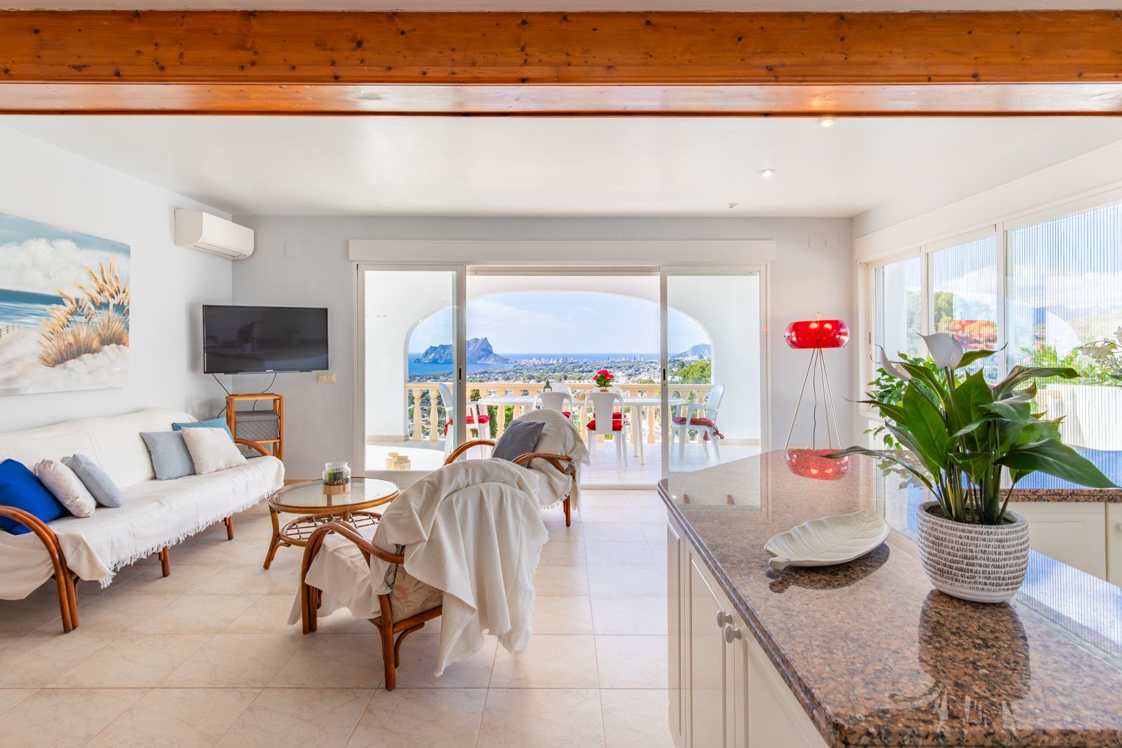 Bright living area featuring exposed wooden beams, granite kitchen island, and sliding doors opening to a terrace with panoramic views of the Peñón de Ifach.