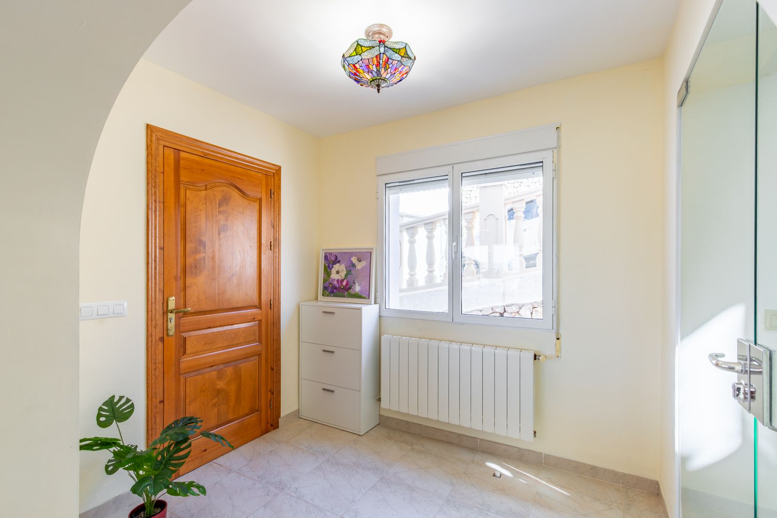 Entrance hallway featuring a solid wood door, central heating radiator, and a Tiffany-style stained glass ceiling light, complemented by neutral tiled flooring.