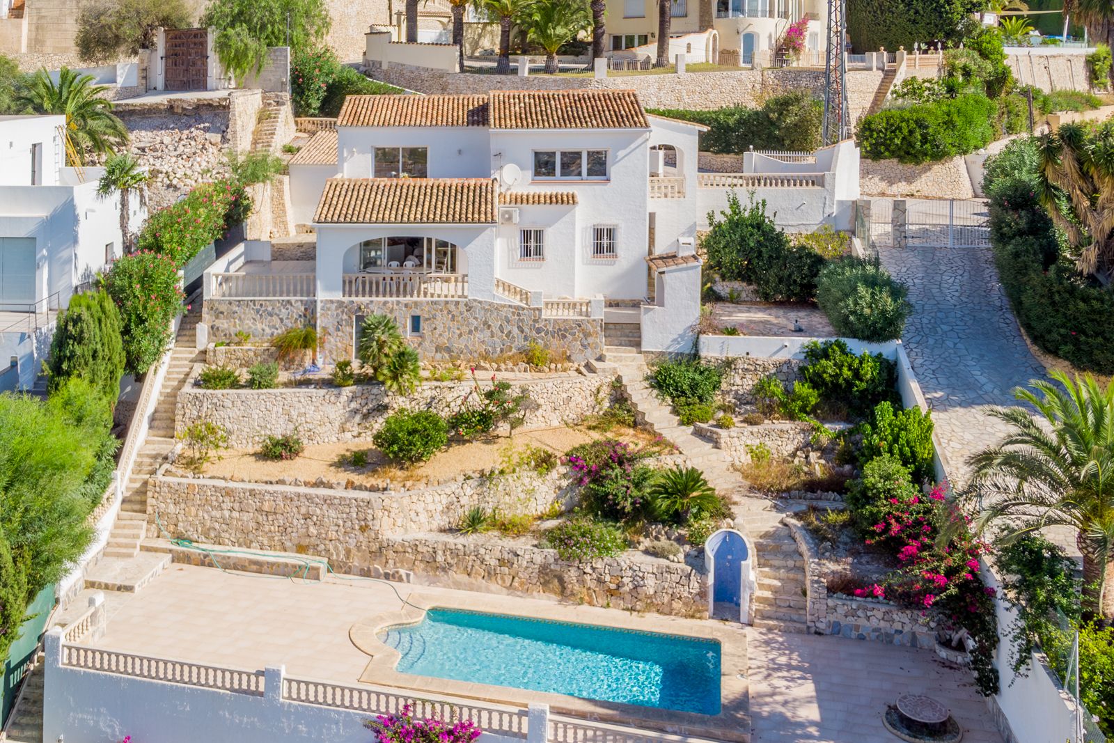Exterior view of the villa featuring white facades, terracotta roofs, and terraced Mediterranean gardens with stone walls leading to the private pool.