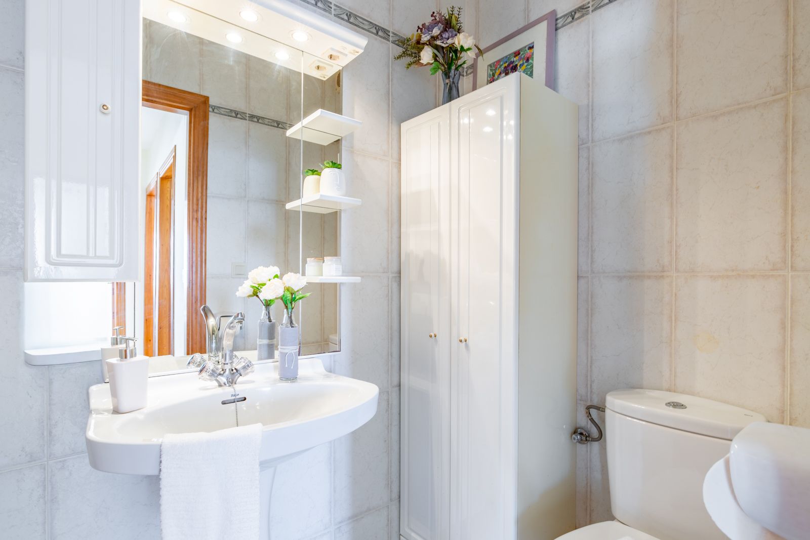 Functional bathroom featuring a pedestal sink, a large mirror with integrated spotlights and glass shelving, and a white storage cabinet against neutral tiling.