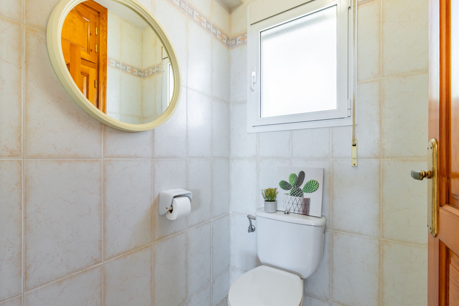 Functional guest toilet featuring beige ceramic wall tiles, a circular mirror with a light frame, and a frosted window for natural light and privacy.