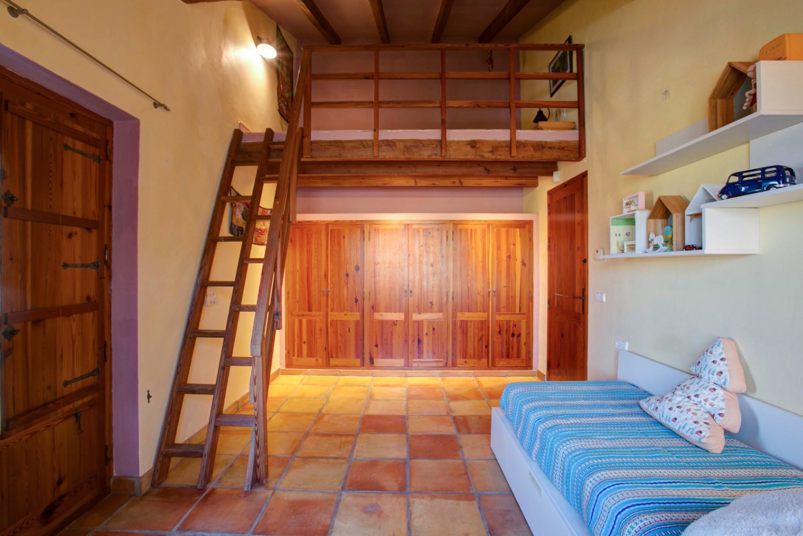 Bedroom featuring a wooden mezzanine with ladder, built-in mobila wood wardrobes, and traditional terracotta flooring under exposed beams.