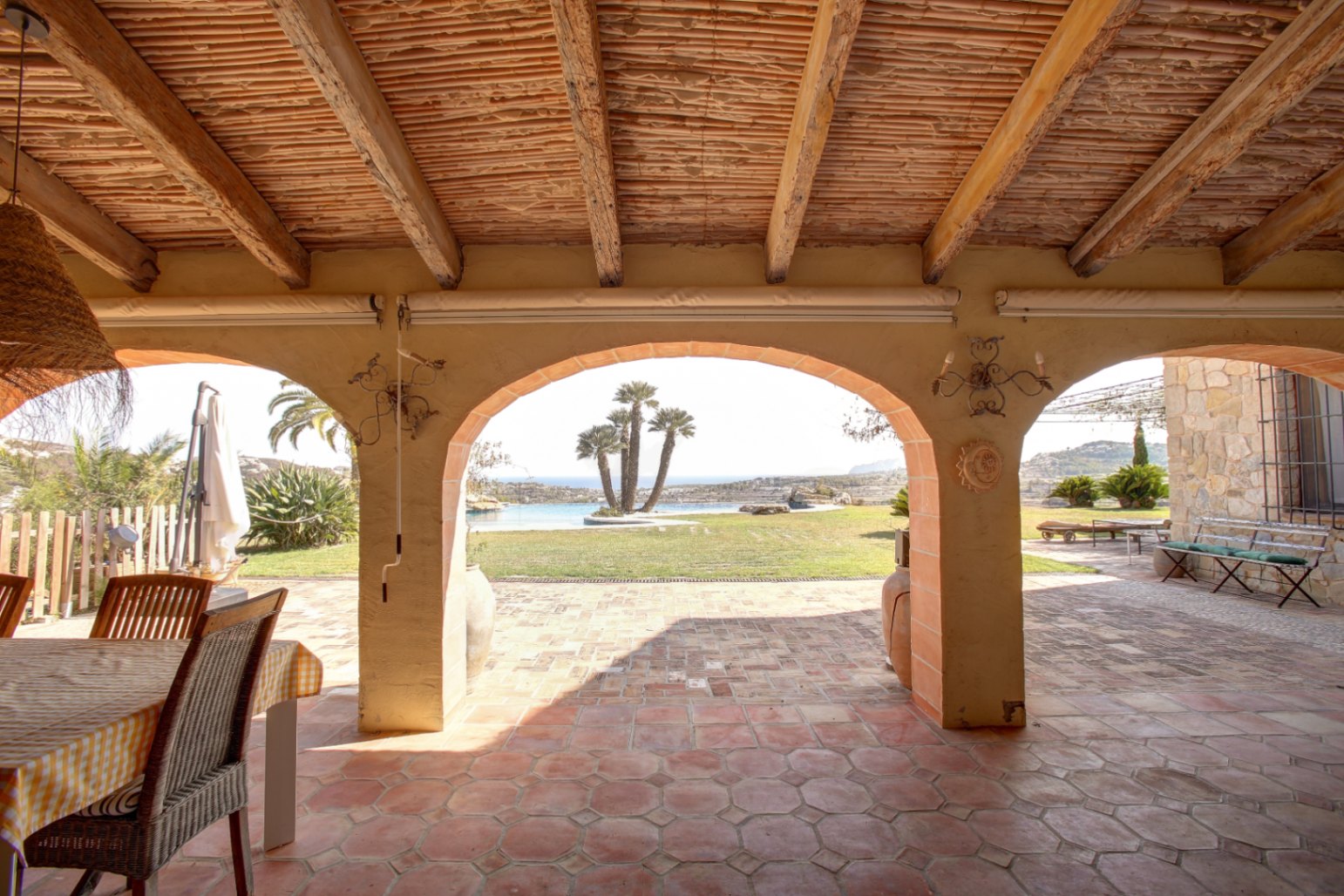 Covered porch featuring exposed wooden beams, reed ceilings, and terracotta flooring. Arched openings frame views of the garden, palm-fringed pool, and Mediterranean Sea.