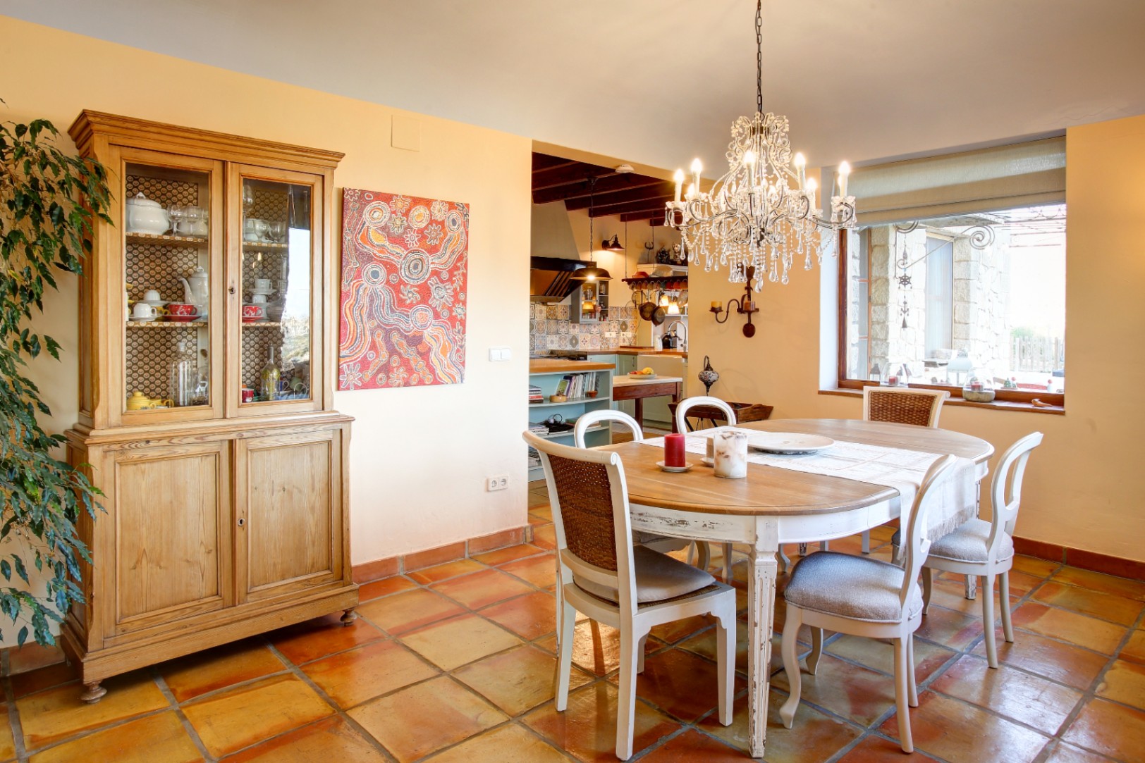 Dining area featuring a wooden table with distressed white chairs, a crystal chandelier, traditional terracotta flooring, and a classic display cabinet.
