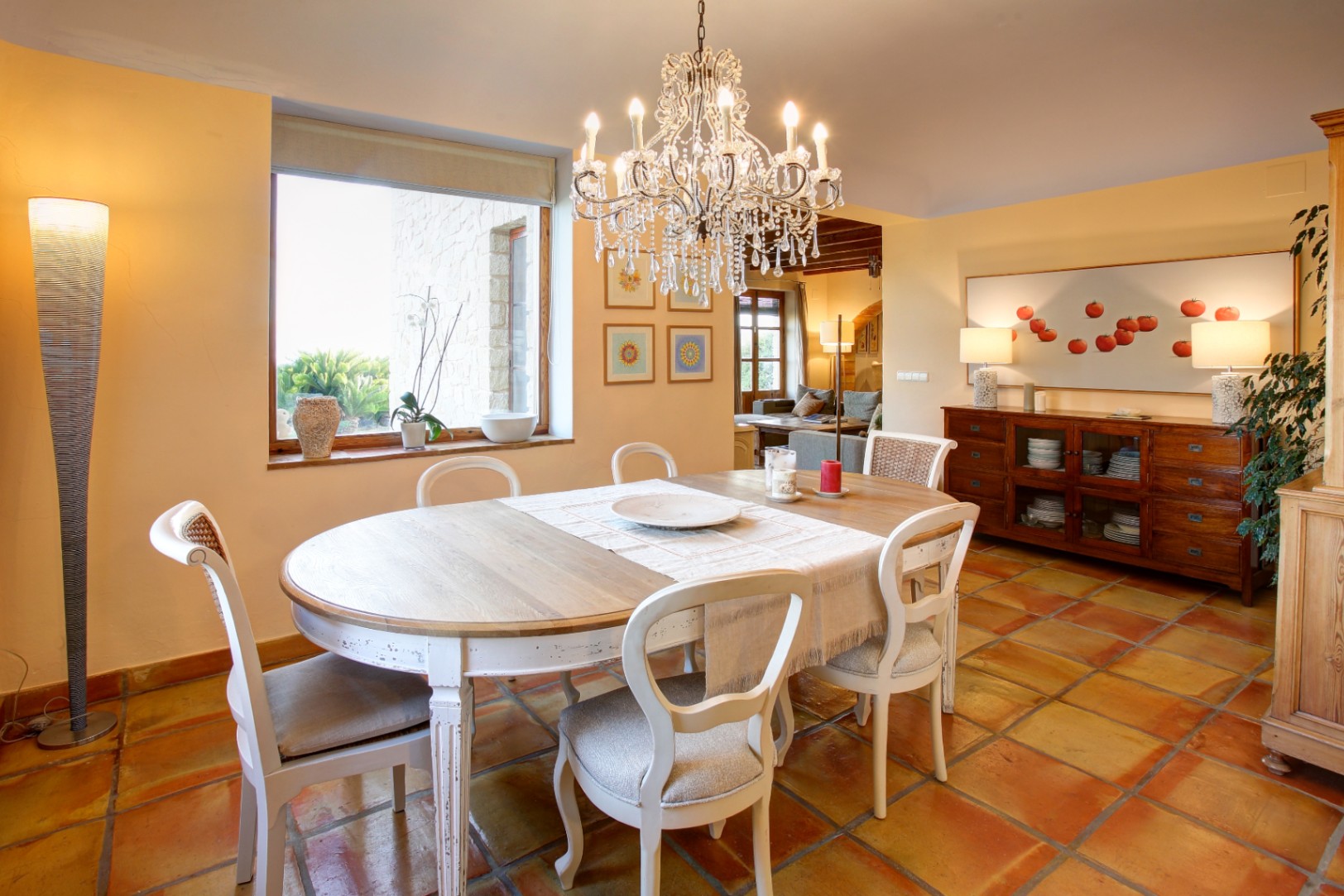 Dining area featuring terracotta floors, a distressed wood table, and a crystal chandelier. Large windows provide natural light and views of the stone exterior walls.